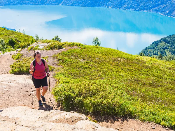 A woman equipped with trekking poles hikes up a mountain trail with a breathtaking view of a blue fjord below.