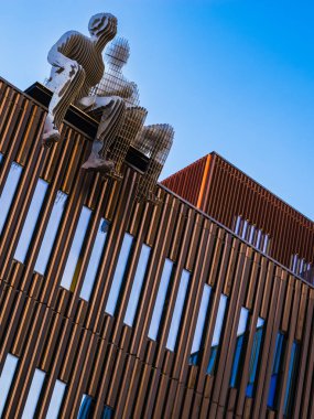 Bu resim, modern bir binanın çatısında, berrak, mavi bir gökyüzünün arka planına karşı duran çarpıcı bir heykeli yakalıyor. Sanat ve mimarinin kombinasyonu yaratıcı ifade ve şehir tasarımının bir karışımını sunar..