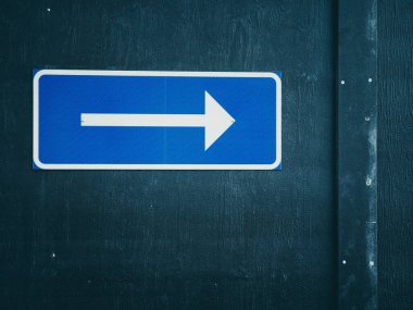 Sağı gösteren beyaz bir oka sahip dikdörtgen mavi bir tabela görünür tahıl ve metal perçin ile koyu ahşap bir duvara monte edilir..