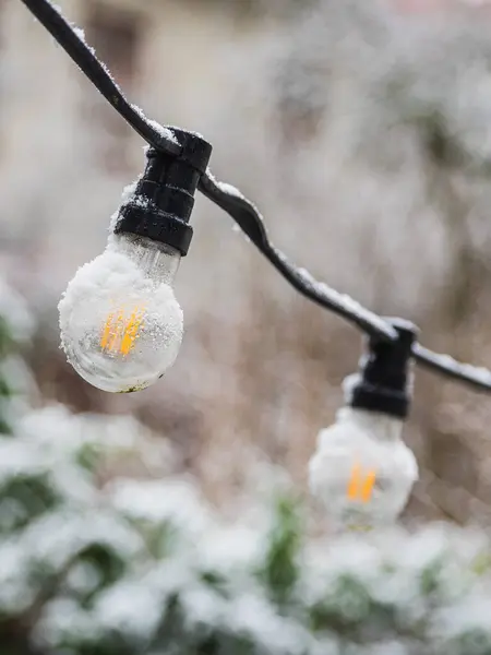 A string of glowing lightbulbs hangs outdoors against a blurred backdrop of white snowfall in Gothenburg, Sweden. Glimpses of snow cling to the bulbs and wire, illuminating a cold winters day with a warm, subtle light.