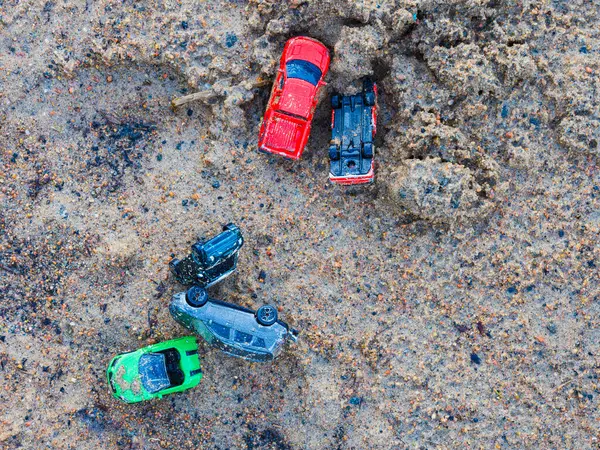 A variety of colorful toy cars is scattered over a gritty, sandy surface, suggesting playful activity. The shot, taken from above, captures the informal arrangement of the toys, possibly following a childs outdoor play session in Sweden.