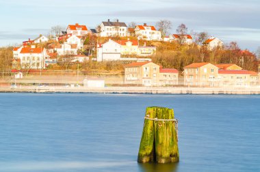 İsveç 'in Göteborg kıyısındaki sakin sularda yosun kaplı bir sütun. Arka planda, İskandinav manzarasını yansıtan, batan güneşin sıcacık parıltısıyla yıkanmış büyüleyici kırmızı çatılı evleri olan sakin bir mahalle yer alıyor..