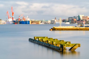 Göteborg Limanı 'nda sakin bir sabah sahnesi. Sakin sulara doğru uzanan yosun kaplı ahşap bir rıhtım. Şehir endüstrisinin arka planı berrak bir gökyüzünün altında yavaşça bulanıklaşıyor..