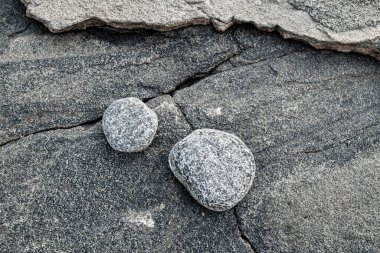 A close-up image of two smooth, grey stones resting on a rocky cliff face. The stones are round and have a textured surface. The cliff face is made of dark grey rock with visible cracks and lines. The image captures the simple beauty of nature and th clipart