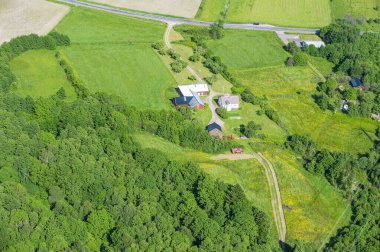 A bird's-eye view of a house in a rural setting. The house is located on a hill, with a large forest to its left and open fields to its right. The house appears to be made of wood and has a red roof. There is also a paved road leading up to the house clipart