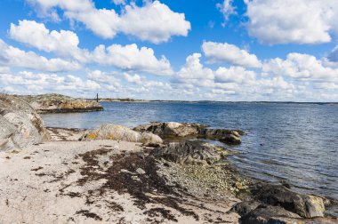 İsveç kıyı şeridinde pürüzsüz kayalar ve deniz yosunlarıyla sakin bir plaj bulunur. Dalgalar kıyı şeridine hafifçe dokunduğunda parlak bulutlar havada süzülür, barışçıl bir atmosfer yaratır..