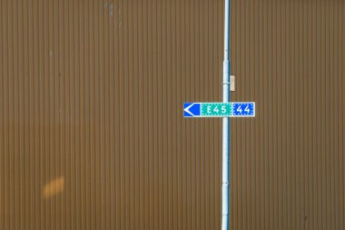 The road sign displays the E45 highway number and road 44, directing traffic to the left, set against a textured brown building backdrop on a clear day. clipart