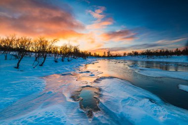 Canlı gündoğumu, gölgeli ağaçların sahneyi çevrelediği karlı bir nehir manzarasının üzerine ılık bir parıltı saçıyor. Su, gökyüzünün turuncu ve mavi renklerini yansıtır..