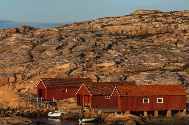 Rustic red wooden houses sit along a rugged coastline, surrounded by smooth rocks and calm waters as the sun sets, casting warm light on the landscape. clipart