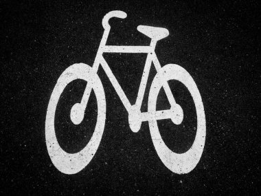 Büyük beyaz bir bisiklet sembolü, hareketli bir kentsel alanda belirlenmiş bisiklet şeridini gösteren koyu asfalta boyanmıştır. Güneş ışığı net tasarımı vurgular.