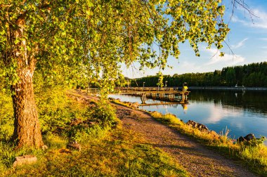 Sakin bir nehir kenarı yolu, canlı yeşil ağaçlar ve batan güneşin sıcacık parıltısıyla aydınlanan barışçıl suyla sarılmış ahşap bir rıhtıma götürür..