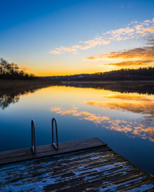 Sakin bir göl, canlı bir gün batımıyla aydınlanır ve suya renkli yansımalar yapar. Adımları olan ahşap bir rıhtım ağaçlarla çevrili sakin bir çevreye açılıyor..