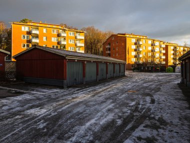 Tamirhaneler yerleşim yerlerinde karlı bir yol çiziyor. Apartman binaları, güneş kara bulutları yarıp geçerken arka planda dik duruyorlar. Sıcak bir ışık yaratıyorlar..