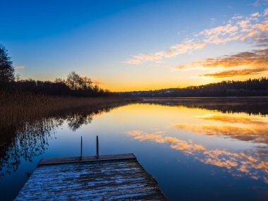 Sakin bir göl, kıyı boyunca ağaçlar tarafından çerçevelenmiş günbatımının parlak renklerini yansıtır. Yumuşak parıltı huzurlu suları aydınlatır ve uyumlu bir atmosfer yaratır..