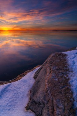 Canlı bir günbatımı gökyüzünde çeşitli renkler bırakır ve kayalık bir sahilin yakınındaki sakin suları yansıtır. Kayalıklarda kar kalır ve dingin manzaraya kış dokunuşu katar..