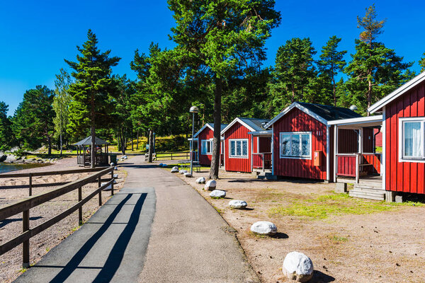 Red wooden cabins line the waterfront, surrounded by lush trees and a well-maintained path. A calm shoreline invites relaxation and exploration on a sunny day in Sweden.