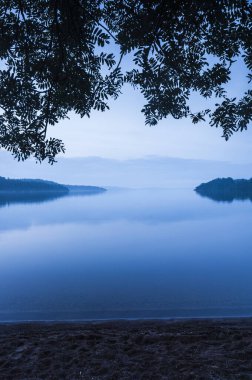 İsveç 'teki Lyngern Gölü' nde sakin, mavi bir sabah görülür. Durgun sular, gökyüzünün yumuşak renklerini yansıtır. Huzurlu bir ortam, bir anlık sessiz bir yansımaya yol açar..