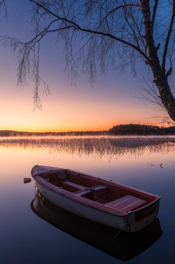 Sakin bir gölde, gün doğumunun yumuşak tonlarını yansıtan küçük bir tekne huzur içinde yüzer. Ağaçların çevresini çevreleyen sahne, İsveç 'in sabahın erken saatlerindeki sakin atmosferini artırıyor..
