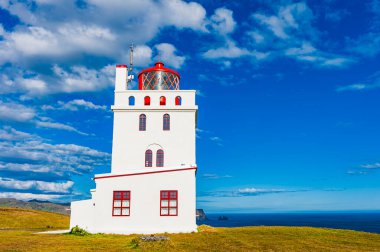 İzlanda 'da, bulutlarla dolu dramatik bir gökyüzü ile çevrili çimenli bir tepenin üzerinde belirgin bir deniz feneri duruyor. Okyanus uzaktan görülebiliyor, manzarayı güzelleştiriyor..