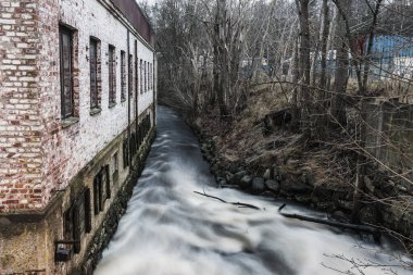 İsveç 'te ağaçların kıyıları kapladığı hafif akan bir nehrin yanında kırsal bir bina duruyor. Sahne, baharın başındaki huzuru yakalıyor, bölgenin doğal güzelliğini vurguluyor..
