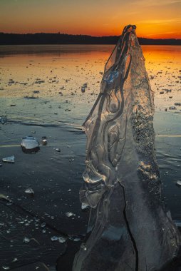 At sunset in Sweden, an impressive ice formation rises from the frozen expanse, reflecting warm hues of gold and orange against a serene background of scattered ice pieces. clipart