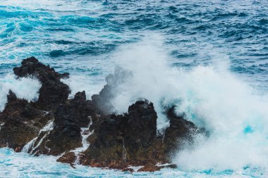 Güçlü dalgalar, fırtınalı bir günde okyanusun ham güzelliğini ve enerjisini gösteren Madeira kıyı şeridi boyunca uzanan karanlık kayalara çarpar. Gökyüzü bulutlu görünüyor, fırtınalı havayı işaret ediyor..