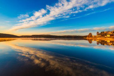 Sabah ışığı sakin bir İsveç gölünü şereflendirir ve sakin suda mükemmel yansımalar yaratır. Yumuşak bulutlar gökyüzünde sürükleniyor ve bu manzaranın huzurlu atmosferini güçlendiriyor..