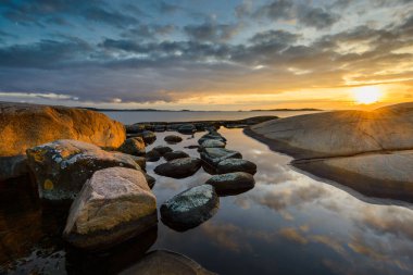 Göteborg yakınlarında gün batımında, kayalık kıyı şeridi sersemletici bir manzara yaratıyor. Gökyüzü renkler ve yansımalarla sakin suyun yüzeyinde dans ederek sükuneti davet ediyor..