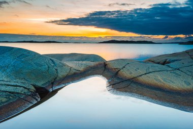 Sakin sular, engebeli kıyı kayaları fışkırırken günbatımının ateşli renklerini yansıtır. Göteborg yakınlarındaki bu sakin yer altın saat boyunca doğanın güzelliğini gözler önüne seriyor..