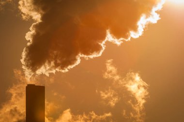 İsveç 'te uzun bir baca, gün batımında havaya yoğun duman salıyor. Gökyüzünün sıcak renkleri karanlık dumanla tezat oluşturarak sanayi faaliyetlerinin çarpıcı bir görüntüsünü oluşturuyor..