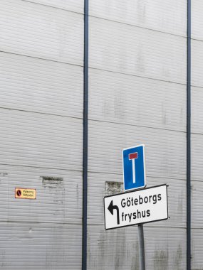 Yol işaretleri Göteborg 'daki soğuk hava deposuna sola dönüş olduğunu gösteriyor. Tabelalar büyük, gri endüstriyel bir yapının önünde duruyor. Basit bir tasarım ve minimal özellikleri var..