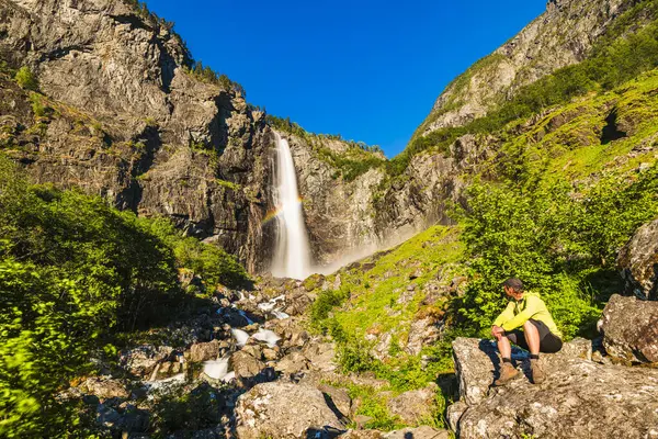 Bir yürüyüşçü, Norveç 'teki Faygefossen şelalesine bakarak bir kayanın üzerinde oturur. Bereketli yeşillik ve kayalık arazi bu açık hava macerasının doğal güzelliğini vurgular..