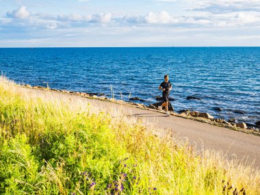 İsveç 'in batı kıyısındaki kıyı şeridi boyunca yürüyen bir erkek koşucu, canlı yeşillik ve parlak bir gökyüzünün altında ışıldayan mavi deniz ile çevrilidir..