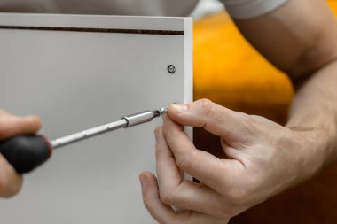 Evde mobilya montajı. Cıvatayı tornavidayla sıkıyorum. Dolap kurulumu. Beyaz tişörtlü bir adam beyaz bir gardırop hazırlıyor. Erkek elleri yakın plan..