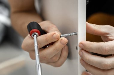 Evde mobilya montajı. Cıvatayı tornavidayla sıkıyorum. Dolap kurulumu. Beyaz tişörtlü bir adam beyaz bir gardırop hazırlıyor. Erkek elleri yakın plan..