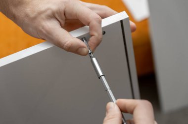 Evde mobilya montajı. Cıvatayı tornavidayla sıkıyorum. Dolap kurulumu. Beyaz tişörtlü bir adam beyaz bir gardırop hazırlıyor. Erkek elleri yakın plan..