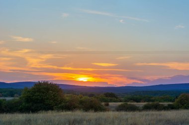 Mavi bulutlu gökyüzüne karşı gün doğumu ya da gün batımı. Bulutlar turuncu ile vurgulanır. Zengin renkler. Seyahat, huzur ve sükunet. Güneş ufukta kayboldu. Dağlar, orman ve ağaçlar