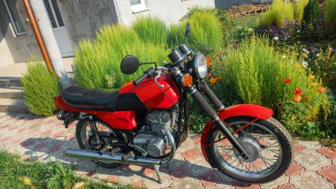 Kırmızı motosiklet Jawa 638 yola park edilmiş. Arka planda ağaçlar ya da çalılar. Yol mesafeye gidiyor. Sonbahar renkleri - sarı, yeşil, kırmızı, mavi. Eski, nadir, koleksiyon motosikletleri.