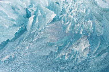 Yakın plan bir buz dokusu kısmen karla kaplı. Yüzeyde, narin çatlaklar ve buz gibi kenarlarla karışık donmuş desenler sergiliyor. Yumuşak, tozlu kar etrafa saçılmış..