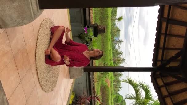 在巴厘岛 年轻美丽的女子穿着红色衣服在废弃的寺庙里沉思和做瑜伽 寺庙里有绿色的手掌和升起的田野背景 女性练习摆姿势 优质Fullhd垂直画面 — 图库视频影像
