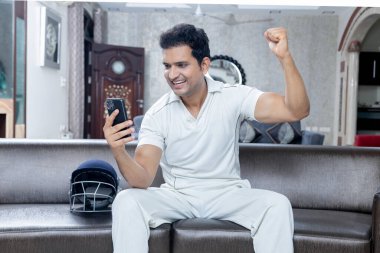 Kriket elbisesi giymiş çok mutlu bir adam oturma odasında koltukta oturup cep telefonuna bakarak kriket izlemekten zevk alıyor.