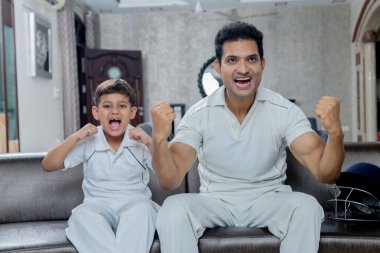 Hintli baba ve oğul kriket elbisesi giyer, kriket seyreder ve evden kriket takımını destekler, baba oğul kriketten zevk alır.