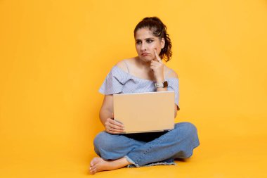 Hintli kız dizüstü bilgisayar kullanarak oturuyor. Renkli arka planda izole edilmiş düşünceli bir ifade.