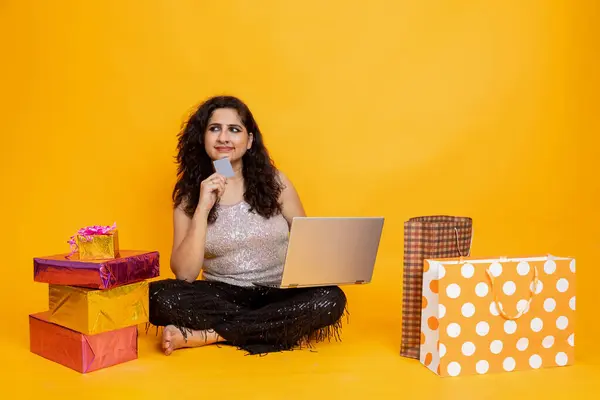 Indian girl sitting on floor with laptop doing online shopping from card, girl with laptop and gift box and shopping bag , online shopping concept