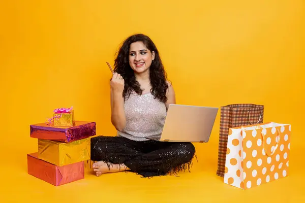 Indian girl sitting on floor with laptop doing online shopping from card, girl with laptop and gift box and shopping bag , online shopping concept