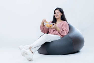 Mutlu Güzel Bayan Fasulye Torbası 'na oturmuş, abur cubur yiyor ve izole edilmiş arka planda filmler izliyor.