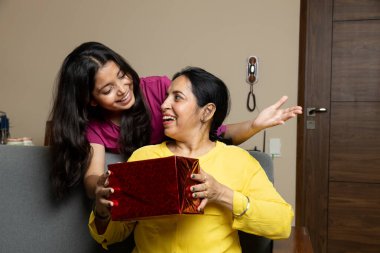 Hintli kız Anneler Günü 'nde anneye sürpriz hediye veriyor, kız ve anne oturma odasında kanepede oturuyor..