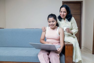 Hintli anne dizüstü bilgisayarla uğraşan kızına cam sütü veriyor. Annesi dizüstü bilgisayarla uğraşırken kızına bakıyor.