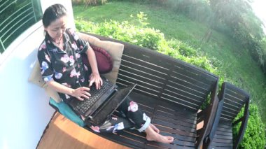 Yaşlı bir Asyalı kadın dizüstü bilgisayarda çalışıyor ve sabahları evin dışındaki sandalyede yatıyor. Uzak çalışma konsepti yüksek kaliteli mobil iletişim ve kablosuz internet.