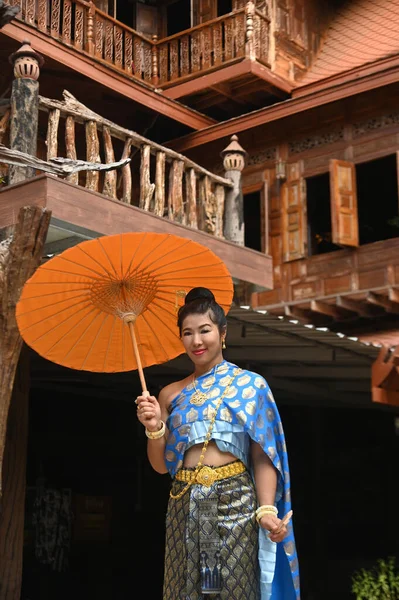 Beautiful Asian woman in Thai National costume holds a paper umbrella posing for photos at a Thai house.